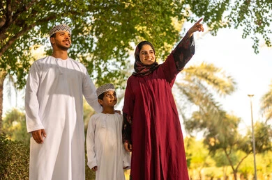 الاستمتاع بالنزهة في الهواء الطلق, تقوية وتعزيز الروابط الأسرية، عائلة عربية خليجية عمانية يرتدون الزي التقليدي يقفون في إحدى الحدائق العامة في النهار, قضاء الوقت الممتع مع الأسرة, حديقة ومنتزه عائلي ترفيهي، الاشارة الي شيء ما