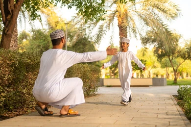 بناء علاقة قويه مع الأولاد، مفهوم الأبوة وحنان الأب، تحمل المسؤولية والاعتناء بالأطفال، المرح وقضاء أوقات ممتعة مع الأبناء فى الحديقة ، صبي عماني عربي خليجي يرتدي الدشداشة و الكمة يقوم بإحتضان والده