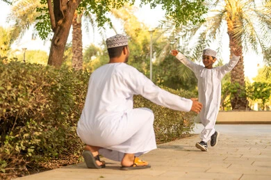 مفهوم الأبوة وحنان الأب، تحمل المسؤولية والاعتناء بالأطفال، المرح وقضاء أوقات ممتعة مع الأبناء فى الحديقة ، صبي عماني عربي خليجي يرتدي الدشداشة و الكمة يقوم بإحتضان والده، بناء علاقة قويه مع الأولاد