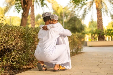 Taking responsibility and caring for children, the concept of fatherhood and a father's affection, having fun and spending enjoyable times with children in the park, an Omani Arab Gulf boy wearing a dishdasha and kumma hugging his father, building a strong relationship with the children.