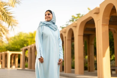 Taking care of one's appearance, going for a walk outdoors in one of the parks and gardens, an Arab Gulf woman from Oman wearing a hijab and abaya looking at something, gestures of happiness and joy, a young Saudi woman standing upright with self-esteem.