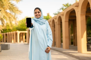 Gestures of happiness and joy, the concept of online shopping, a Saudi Gulf Arab woman displaying a blank black screen, taking a stroll outdoors in one of the parks and recreational areas, a young Omani woman wearing an abaya and hijab holding a mobile phone in her hands.