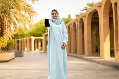 The concept of online shopping, a Saudi Gulf Arab woman displaying a blank black screen, taking a stroll outdoors in one of the parks and recreational areas, an Omani young woman wearing an abaya and hijab holding a mobile phone in her hands, gestures of happiness and joy.
