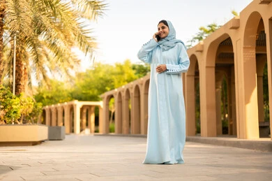 Chatting with family and friends, an Omani woman wearing an abaya and hijab is making a phone call via her mobile, gestures and expressions of happiness and joy, a young Arab Gulf Saudi woman using a mobile phone, using modern technology, taking a stroll outdoors in one of the parks and recreational areas.