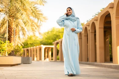 Taking a stroll outdoors in one of the parks and gardens, an Omani woman wearing an abaya and hijab is making a phone call on her mobile, with gestures and expressions of happiness and joy. A young Saudi Gulf Arab woman is using a mobile phone, utilizing modern technology, and enjoying pleasant times.