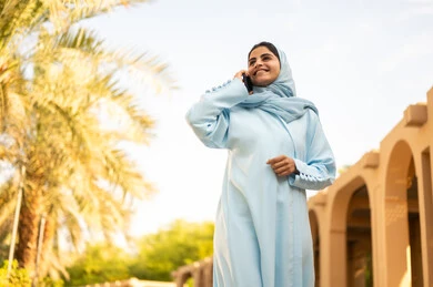 شابة عربية خليجية سعودية تستخدم الهاتف المحمول تتجول في إحدى الحدائق العامة، استخدام جهاز تقني حديث، قضاء أوقات ممتعة، ايماءات وتعابير السعادة و السرور، إمرأة عمانية ترتدي العباءة والحجاب تقوم باجراء مكالمه هاتفية عبر الجوال،  القيام بنزهه في الهواء الطلق في احدى الحدائق والمنتزهات