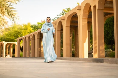 Strolling and walking in green parks, having a picnic outdoors and relaxing, a Saudi Arabian Gulf woman practicing walking, enjoying natural parks, a young Omani woman in a hijab wearing an abaya is taking a walk in the garden.