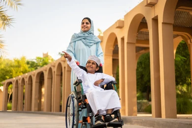قضاء لحظات سعيدة مع الأبناء أثناء العطلة، عائلة عربية عمانية خليجية في رحلة بإحدى الحدائق في سلطنه عمان ، ولد عربي خليجي عماني يرتدي الدشداشة و الكمه يجلس على كرسي متحرك يقوم بالاشارة الي شيء ما برفقه والدته ، ايماءات السعادة وا لسرور