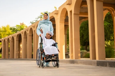 Spending enjoyable and fun time with children during the summer vacation, an Arab Omani Gulf family on a trip to one of the parks in the Sultanate of Oman. An Arab Gulf Omani boy wearing a dishdasha and a kumma sits in a wheelchair, pointing to something with his mother, gestures of happiness and joy.