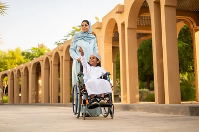الأنشطة الترفيهية مع الأبناء أثناء الإجازة، عائلة عربية عمانية خليجية في رحلة بإحدى الحدائق في سلطنه عمان ، ولد عربي خليجي عماني يرتدي الدشداشة و الكمه يجلس على كرسي متحرك يقوم بالاشارة الي شيء ما برفقه والدته ، ايماءات السعادة وا لسرور
