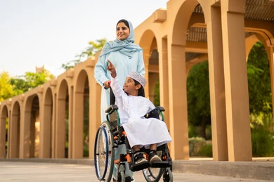 مشاركة الأبناء لحظات ممتعة في العطلة، عائلة عربية عمانية خليجية في رحلة بإحدى الحدائق في سلطنه عمان ، ولد عربي خليجي عماني يرتدي الدشداشة و الكمه يجلس على كرسي متحرك يقوم بالاشارة الي شيء ما برفقه والدته ، ايماءات السعادة وا لسرور