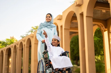 قضاء وقت ممتع مع الأبناء في الإجازة، عائلة عربية عمانية خليجية في رحلة بإحدى الحدائق في سلطنه عمان ، ولد عربي خليجي عماني يرتدي الدشداشة و الكمه يجلس على كرسي متحرك يقوم بالاشارة الي شيء ما برفقه والدته ، ايماءات السعادة وا لسرور