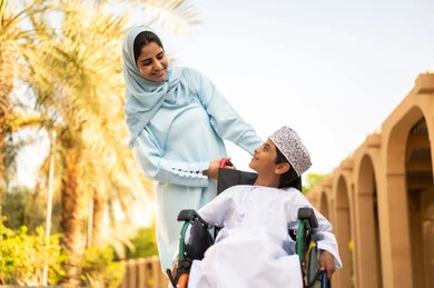 An Arab Omani Gulf family on a trip to one of the parks in the Sultanate of Oman, the concept of a happy family, spending enjoyable family time, cheerful family atmosphere, weekend, an Arab Gulf Omani boy wearing a dishdasha and a kumma sitting in a wheelchair looking at his mother with gestures of love and happiness.