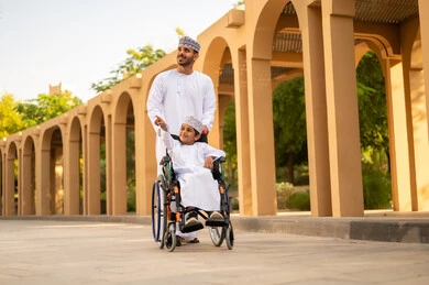 الطبيعة الخضراء في سلطنة عمان، تمضية أوقات ممتعة مع العائلة في الحديقة، ولد عربي خليجي عماني يرتدي الدشداشة و الكمه يجلس على كرسي متحرك يقوم بالاشارة الى شيء ما، اب يقوم بقضاء وقت ممتع مع ابنه فى الخارج الطبيعة الخضراء في سلطنة عمان، تمضية أوقات ممتعة مع العائلة في الحديقة، ولد عربي خليجي عماني يرتدي الدشداشة و الكمه يجلس على كرسي متحرك يقوم بالاشارة الى شيء ما، اب يقوم بقضاء وقت ممتع مع ابنه فى الخارج