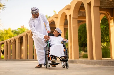 ولد عربي خليجي عماني يرتدي الدشداشة و الكمه يجلس على كرسي متحرك ينظر الى ابيه بايماءات الحب و الفرح، حب الآباء لأبنائهم, مفهوم الأسرة السعيدة ، مشاركة الأبناء لحظات مرحة في الحديقة