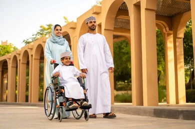 The view of green grass and trees in broad daylight, family conversations and discussions in the open air, an Omani Gulf Arab couple dressed in traditional attire walking and strolling in the public park accompanied by their son sitting in a wheelchair, the concept of a happy family.