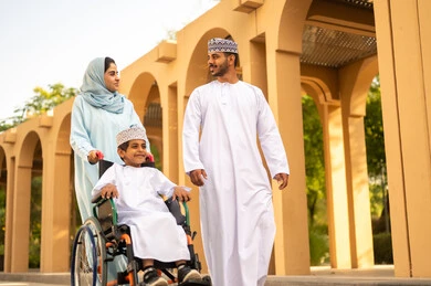 الحوارات والنقاشات العائلية في الهواء الطلق، منظر العشب والأشجار الخضراء في وضح النهار، زوجان عربيان خليجيان عمانيان يرتديان الزي التقليدي يقومان بالمشي والتنزه في الحديقة العامة برفقة ابنهم يجلس على كرسي متحرك، مفهوم الاسرة السعيدة