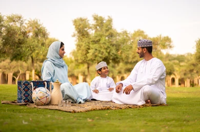 Strengthening and enhancing family bonds, the concept of a happy family, an Arab Gulf Omani family dressed in traditional attire sitting in an outdoor park, exchanging conversations with gestures of joy.