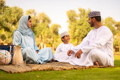قضاء عطله نهاية الاسبوع فى الخارج،اللعب والتسلية مع الأبناء في الحديقة ، مفهوم الاسرة السعيدة، عائله عربية خليجيه عمانية ترتدي الزي التقليدي تجلس فى احدي الحدائق فى الهواء الطلق، ايماءات السعادة و السرور