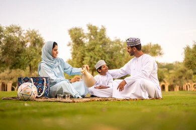 Sharing joyful moments with children in the park, enjoying drinks, the concept of a happy family, an Omani Gulf Arab family dressed in traditional attire sitting in an outdoor park, gestures of happiness and joy, spending the weekend outdoors.