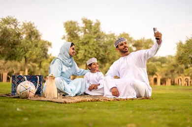 مفهوم الاسرة السعيدة، التقاط صور تذكارية عبر الهاتف المحمول ، عائله عربية خليجيه عمانية ترتدي الزي التقليدي تجلس فى احدي الحدائق فى الهواء الطلق، ايماءات السعادة و السرور، قضاء عطله نهاية الاسبوع فى الخارج