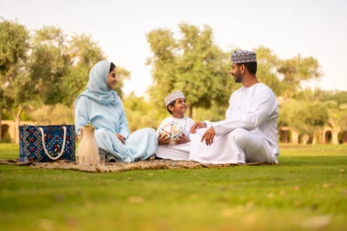 An Omani Gulf Arab family dressed in traditional attire is spending enjoyable time outdoors, playing and having fun with their children in the park, enjoying engaging in youth activities and sports. An Omani Gulf Arab boy is holding a football sitting with his parents, showing gestures of happiness and joy.