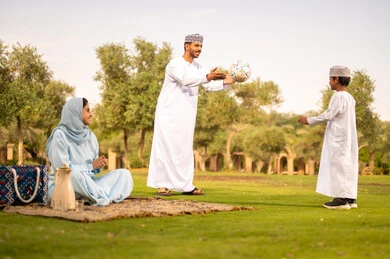 مشاركة الأبناء لحظات مرحة في الحديقة، مفهوم اللياقة والرشاقة البدنية، الاستمتاع بممارسة الأنشطة والرياضات الشبابية، رجل عربي خليجي عماني يرتدي الدشداشة والكمة يقوم بلعب كرة القدم مع ابنه ، عائله عربية خليجيه عمانية ترتدي الزي التقليدي تقوم بقضاء وقت ممتع فى الخارج ، ايماءات السعادة و السرور