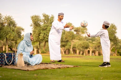 Spending enjoyable times with children in the park, the concept of fitness and physical agility, enjoying engaging in youth activities and sports, an Arab Gulf Omani man wearing a dishdasha and kumma playing football with his son, a father encouraging his son with gestures of happiness and joy.