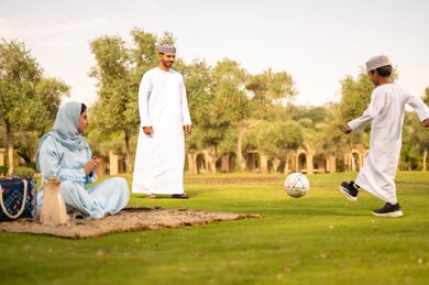 Enjoying engaging in youth activities and sports, the concept of fitness and physical agility, an Arab Gulf Omani man wearing a dishdasha and kumma playing football with his son, a father encouraging his son with gestures of happiness and joy, spending enjoyable times with children in the park.