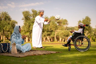 Enjoying sports and physical activities, spending enjoyable times with children in the park, the concept of fitness and physical agility, an Arab Gulf Omani boy with special needs wearing a dishdasha and a kumma sitting in a wheelchair playing ball with his father, a father encouraging his son with gestures of happiness and joy.