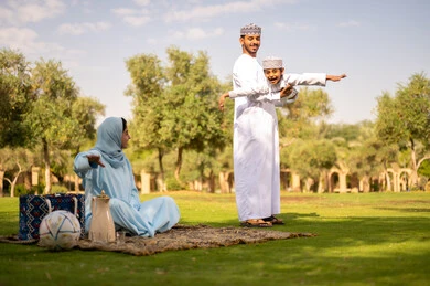 Family bonding and compassion, an Arab Gulf Omani family dressed in traditional attire enjoying the outdoors, an Arab Gulf Omani man wearing a dishdasha and kumma lifting his son up high, gestures of happiness and joy.
