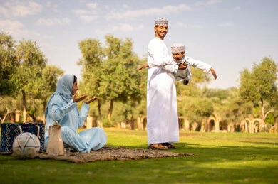 عطلة نهاية الأسبوع ،أجواء عائلية مرحة, عائله عربيه خليجيه عمانية ترتدي الزي التقليدي تقوم بالاستمتاع  بالهواء الطلق، رجل عربي خليجي عماني يرتدي الدشداشة والكمة يقوم برفع ابنه للاعلي ، إيماءات السعادة والفرح