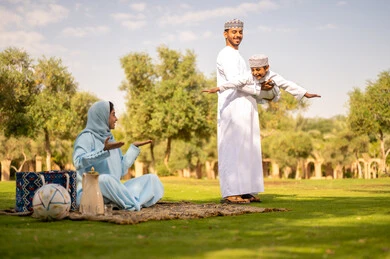 أجواء عائلية مرحة, عائله عربيه خليجيه عمانية ترتدي الزي التقليدي تقوم بالاستمتاع  بالهواء الطلق، رجل عربي خليجي عماني يرتدي الدشداشة والكمة يقوم برفع ابنه للاعلي ، إيماءات السعادة والفرح