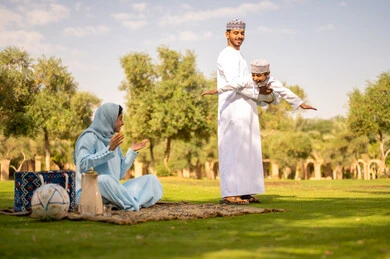 حديقة ومنتزه ترفيهي عائلي, عائله عربيه خليجيه عمانية ترتدي الزي التقليدي تقوم بالاستمتاع  بالهواء الطلق، رجل عربي خليجي عماني يرتدي الدشداشة والكمة يقوم برفع ابنه للاعلي ، إيماءات السعادة والفرح