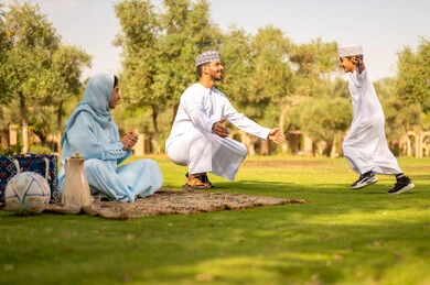 قضاء أوقات عائلية ممتعة بالعطلة الصيفية في الحديقة، عناق الأبناء لأبائهم، حب الآباء لأبنائهم، أب عربي خليجي عماني يرتدي الدشداشة و الكمة يعانق إبنه بحنان, مفهوم الأسرة السعيدة , ايماءات طفل تدل على الحب و الفرح