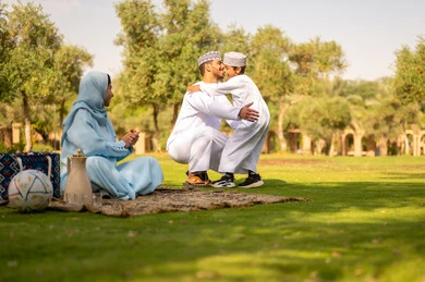 عناق الأبناء لأبائهم، حب الآباء لأبنائهم، أب عربي خليجي عماني يرتدي الدشداشة و الكمة يعانق إبنه بحنان, مفهوم الأسرة السعيدة ، قضاء أوقات عائلية ممتعة بالعطلة الصيفية في الحديقة, ايماءات طفل تدل على الحب و الفرح