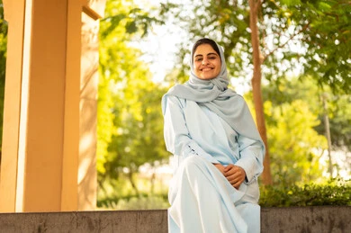 الاستمتاع بالجلوس في الطبيعة الخضراء في الهواء الطلق، سيده عمانية تقوم بنشاط ترفيهي مسلي، قضاء وقت ممتع في الحديقة العامة، فتاة عربية خليجية سعودية مبتسمة ترتدي الحجاب والعباءة تنظر إلى الكاميرا  بإيماءات السرور والسعادة