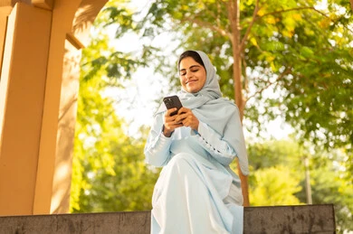 The use of mobile phones to communicate with individuals, chatting, conversations, and making acquaintances. A Gulf Arab Saudi woman wearing an abaya and hijab sits outdoors in one of the parks. An Omani woman browses her phone, showing gestures of happiness and joy.