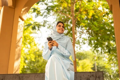 سيده عمانية تتصفح الجوال، متابعة أخبار السوشال ميديا ومواقع التواصل الإجتماعي، استخدام الهاتف المحمول في التواصل مع الأفراد، تصوير من الجانب لامراة عربيه خليجيه سعودية ترتدي العباءة و الحجاب تجلس فى الهواء الطلق فى احدي الحدائق و تنظر الى شيء ما، ايماءات السعادة و السرور