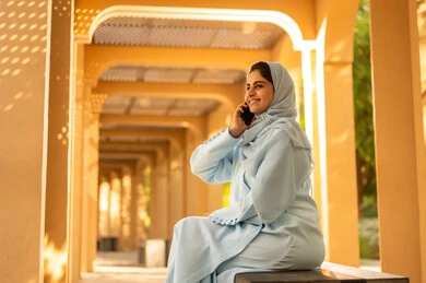 The concept of maintaining family ties, an Omani woman using modern and advanced devices and technologies, exchanging entertaining conversations via mobile, communicating and checking on friends and relatives, a side shot of an Arab Gulf Saudi woman wearing an abaya and hijab sitting outdoors in one of the parks making a phone call.