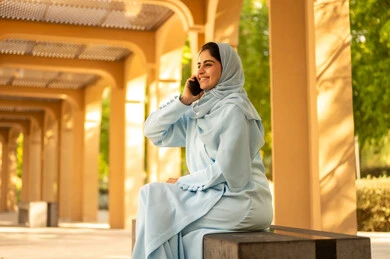 Exchanging entertaining conversations via mobile, communicating and checking on friends and relatives, a side shot of a Saudi Gulf Arab woman wearing an abaya and hijab sitting outdoors in a park making a phone call, an Omani woman using modern and advanced devices and technologies, the concept of maintaining family ties.