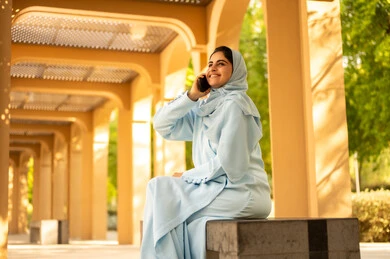 التواصل و الاطمئنان على الأصدقاء والأقارب، تبادل الأحاديث المسليه عبر الجوال، تصوير من الجانب لإمرأة عربية خليجية سعودية ترتدي العباءة والحجاب تجلس فى الهواء الطلق فى احدي الحدائق تقوم باجراء مكالمة هاتفية، سيدة عمانية تقوم باستخدام الأجهزة والتقنيات الحديثة والمتطورة، مفهوم صلة الرحم