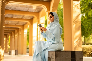 Following news on social media and social networking sites, using mobile phones to communicate with individuals, a side shot of a Gulf Arab Saudi woman wearing an abaya and hijab sitting outdoors in one of the parks, an Omani woman browsing her phone, gestures of happiness and joy.