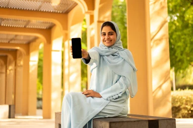 قضاء وقت مسلى وممتع خارج المنزل، استخدام الوسائل التقنية الحديثة، شابة عمانية ترتدي الحجاب و العباءة تقوم بعرض شاشة سوداء فارغة، امراة عربيه خليجيه سعودية تجلس فى احدي الحدائق تمسك بيدها الهاتف المحمول، ايماءات السعادة و السرور