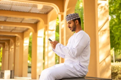 التسوق الالكتروني وتصفح برامج وتطبيقات الجوال، تصوير من الجانب رجل عربي خليجي عماني يرتدي الدشداشة و الكمة يجلس فى الهواء الطلق يمسك بيدة الهاتف المحمول، ايماءات السعادة و السرور