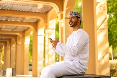 تطور التكنولوجيا والأجهزة التقنية، الدردشة مع الأهل والأصدقاء، تصوير من الجانب رجل عربي خليجي عماني يرتدي الدشداشة و الكمة يجلس فى الهواء الطلق يمسك بيدة الهاتف المحمول و ينظر الي شيء ما، ايماءات السعادة و السرور