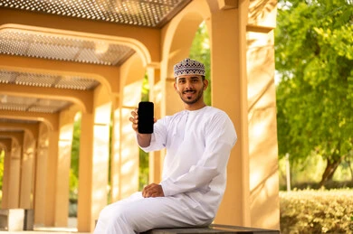 الاستمتاع بقضاء وقت ممتع فى الخارج، استخدام الوسائل التقنية الحديثة، عرض شاشة سوداء فارغة، رجل عربي خليجي عماني يرتدي الدشداشة و الكمة يجلس فى احدي الحدائق يمسك بيدة الهاتف المحمول، ايماءات السعادة و السرور
