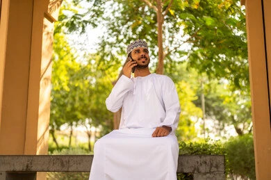 ايماءات السعادة لسماع الاخبار المبهجة ، برامج وتطبيقات الجوال المتنوعة ، اجراء مكالمة هاتفية عبر الجوال ، الاطمئنان على الأصدقاء، تبادل الاحاديث والاخبار بين العائلة، رجل عربي خليجي عماني يرتدي الدشداشة و الكمه يجلس فى احد الحدائق يمسك بيده الهاتف المحمول، النظر الي شيء ما