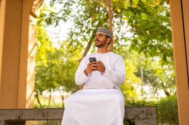 استخدام الهاتف المحمول في التواصل مع الأفراد، دردشة ومحادثات وتعارف، رجل عربي خليجي عماني يرتدي الدشداشة و الكمة يجلس فى الهواء الطلق يمسك بيدة الجوال و ينظر الى شيء ما ، ايماءات السعادة و السرور