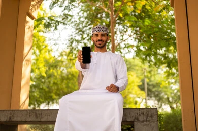 Admiring mobile phone applications, using modern devices and technologies, displaying a blank black screen, an Arab Gulf Omani man wearing a dishdasha and kumma sitting outdoors holding a mobile phone in his hand, gestures of happiness and joy.